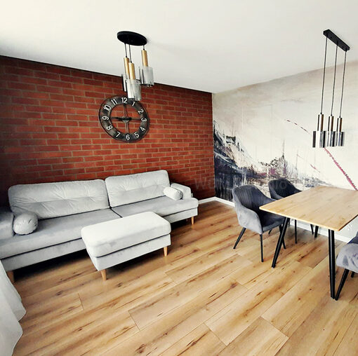 Zdjęcie salonu w stylu loftowym. Po lewej stronie widać szarą sofę z podnóżkiem. Za nią cała ściana pokryta jest cegłą. Na środku ściany duży zegar ścienny. Po prawej stronie stół drewniany z czarnymi nogami oraz cztery krzesła szare welurowe. Nad nim trzy lampy wiszące, pasujące do plafonu, który znajduje się w strefie sofy. Stół stoi przy ścianie z ozdobną tapetą. Na ścianie drzwi ukryte prowadzące do garderoby.