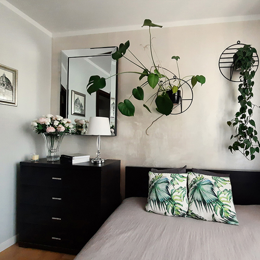 Zdjęcie sypialni w stylu glamour w mieszkaniu. Na pierwszym planie łóżko podwójne z poduszkami dekoracyjnymi. Nad łóżkiem dwa nowoczesne kwietniki ścienne z roślinami zielonymi. Po lewej stronie łóżka komoda z czterema szufladami, lampą stojącą oraz kryształowym wazonem z bukietem kwiatów. Nad komodą duże lustro w ramie lustrzanej, w której odbija się obraz wiszący na ścianie obok.