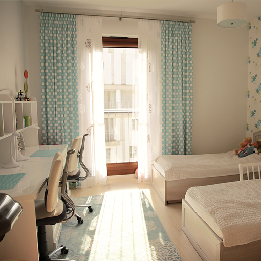 Zdjęcie pokoju dziecięcego. Po prawej stronie we wnęce dwa łóżka dziecięce w kolorze dębu bielonego. Z tyłu na ścianie tapeta dziecięce z kolorowymi kwiatami. Po lewej stronie dwa biurka białe razem z nadbudówkami oraz dwoma krzesłami biurowymi. Na wprost zdjęcia okno z firankami i zasłonkami turkusowymi z białymi kropkami oraz dywan turkusowy na podłodze.